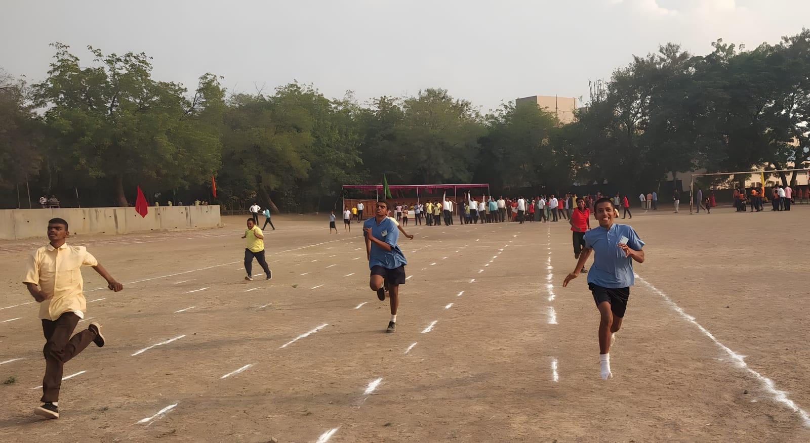 Students competing in a running race on a marked track, with spectators watching from the far side of the ground